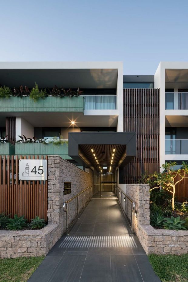 Street-facing exterior view of a contemporary multi-storey residential building entrance. The facade combines white rendered walls, dark vertical timber screening, green-tinted glass balcony balustrades, stone retaining walls, and landscaped planting. A covered, well-lit accessible walkway leads to the recessed entry, with the building number 45 displayed beside the entrance.