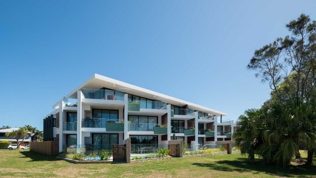 Exterior view of a contemporary multi-storey residential apartment building with a long, shallow pitched or sloping roof, white rendered structural frames, extensive glass balcony balustrades, dark vertical cladding elements, and landscaped lawn and palms in the foreground.