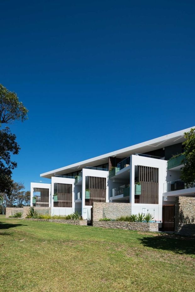 Exterior view of a contemporary multi-unit residential building with a long, low-slung roofline, white rendered walls, projecting balconies, vertical timber or metal screening, and lower stone-clad walls. The building sits behind a landscaped lawn with mature trees under a clear blue sky.
