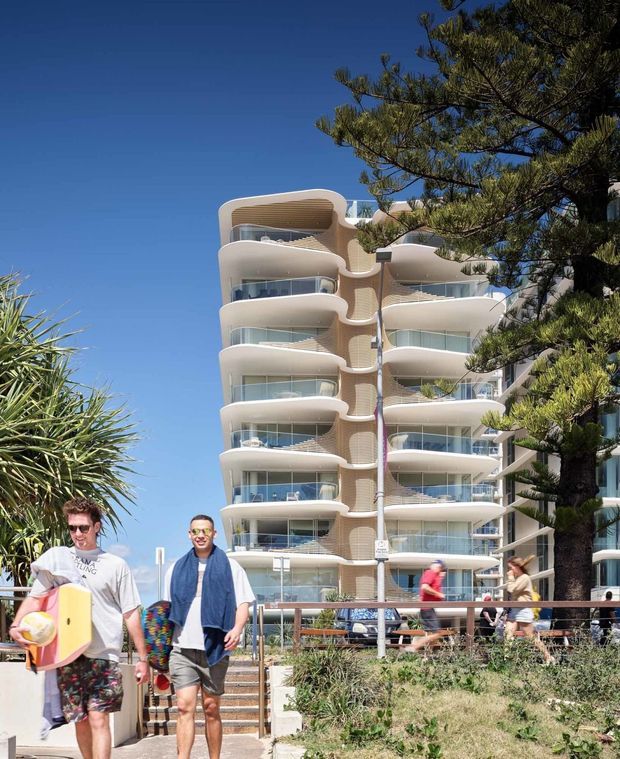 Bright exterior street-level view of a contemporary multi-storey apartment building with rounded, sculptural balconies, curved glass balustrades, and a light-coloured facade. Pedestrians and landscaped coastal-style vegetation occupy the foreground, with a tall evergreen tree partially obscuring the right side. Clear blue sky and strong sunlight create a distinctly coastal urban setting.