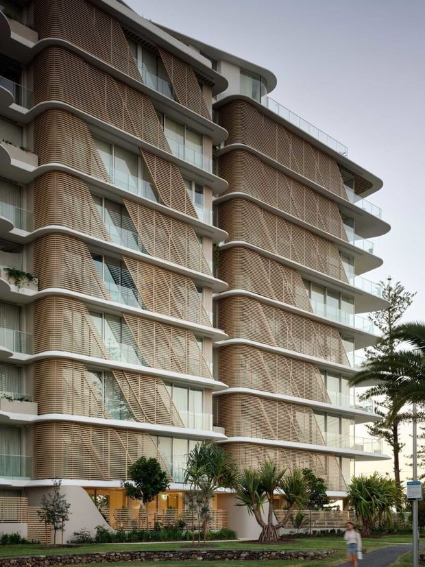 Street-level exterior view of a multi-storey contemporary apartment building with strongly rounded corners, projecting floor slabs, extensive glazing, and repeating curved vertical screens made from horizontal timber-toned louvers. Landscaped planting and a pedestrian path sit along the base, with balconies and rooftop railings visible.