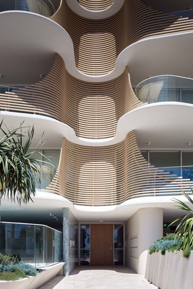 A multi-storey contemporary building entrance and atrium with dramatic curved balconies, white rendered surfaces, glass balustrades, and repeated horizontal timber-look slats forming sculptural wave-like screens. The ground-level entry features a timber door, pale paving, curved glazing, cylindrical columns, and integrated planters with drought-tolerant landscaping.