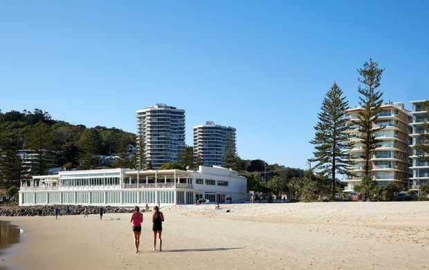 Wide beach scene showing a low-rise beachfront building with extensive glazing and a white facade, backed by several modern multi-storey apartment towers and landscaped coastal vegetation. Two people walk across the sandy foreground under a clear blue sky.