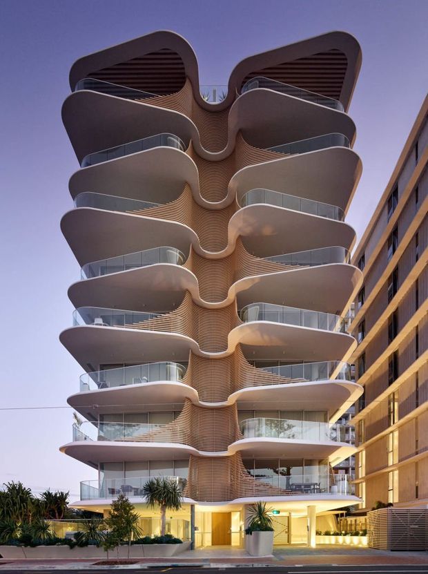 A striking multi-storey residential apartment building photographed from the street at dusk. The facade features repeated curving balconies with glass balustrades, rounded projecting slabs, and a central ribbed vertical feature that creates an organic sculptural form. Warmly lit ground-floor entry landscaping and a neighbouring apartment building are visible.