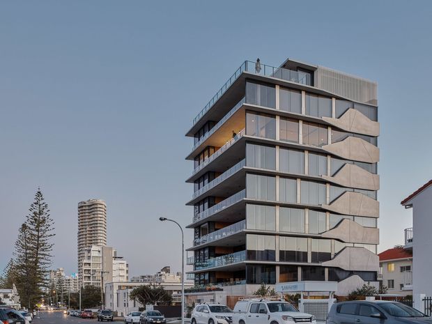 Street-level exterior photograph of a contemporary multi-storey apartment building with stacked projecting balconies, extensive floor-to-ceiling glazing, dark horizontal slab edges, and pale textured facade panels. Urban surroundings include neighbouring buildings, parked cars, and mature street trees.
