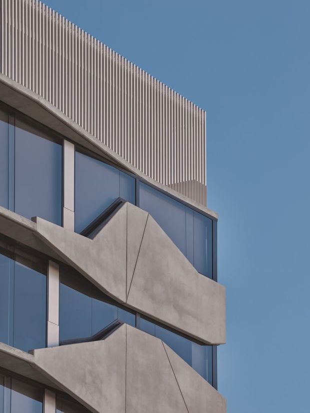 Close-up architectural view of a contemporary multi-storey building facade with exposed concrete projecting balcony forms, expansive blue-tinted glazing, and a rooftop screen of closely spaced vertical fins against a clear blue sky.