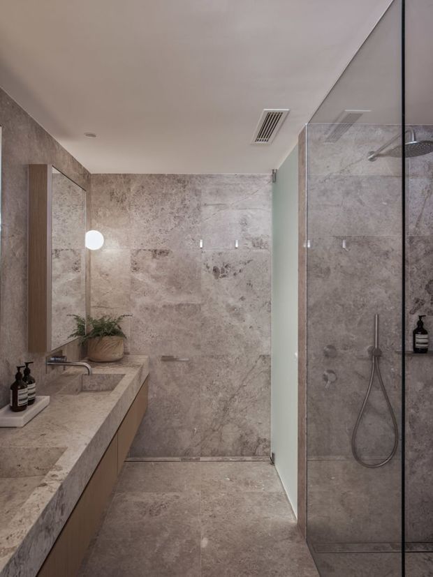 Contemporary bathroom with a walk-in glass shower, large-format beige-grey stone-look wall and floor tiling, a stone vanity benchtop with integrated basin, warm wood-toned cabinetry, brushed metal fixtures, and understated greenery.