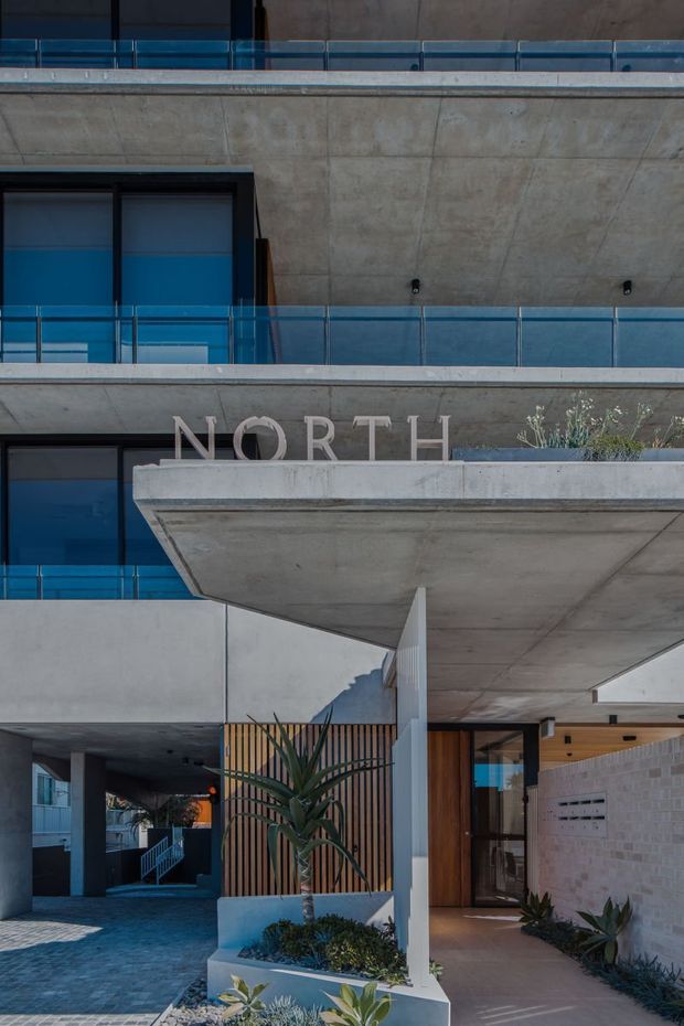 Contemporary multi-storey residential building facade with exposed concrete slabs and walls, blue-tinted glazing, transparent glass balcony balustrades, and a projecting concrete entry canopy. The ground-level entrance combines vertical timber screening, a timber door, pale masonry, integrated planters, and drought-tolerant landscaping. The building signage reads “NORTH.”
