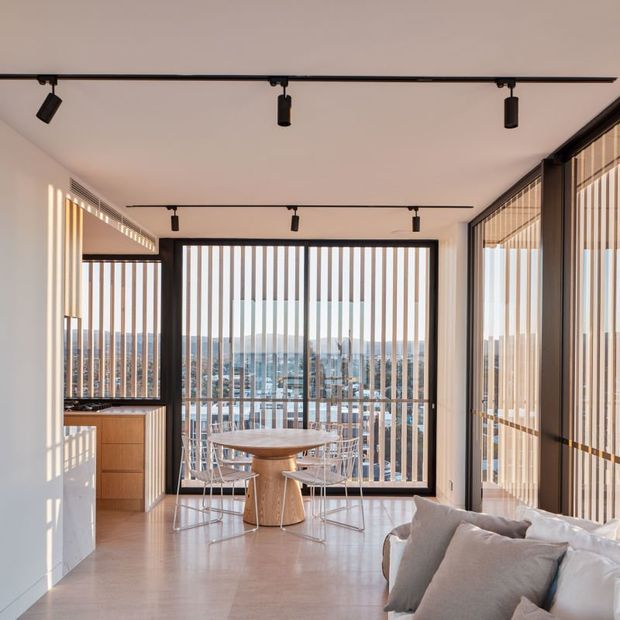 Bright contemporary apartment interior with an open-plan dining and living area overlooking a cityscape. Full-height glazing is screened by vertical timber-look slats, with black-framed windows, track lighting, pale flooring, a compact timber kitchen, a round dining table, and a light neutral sofa. Strong sunlight creates a warm, airy atmosphere.