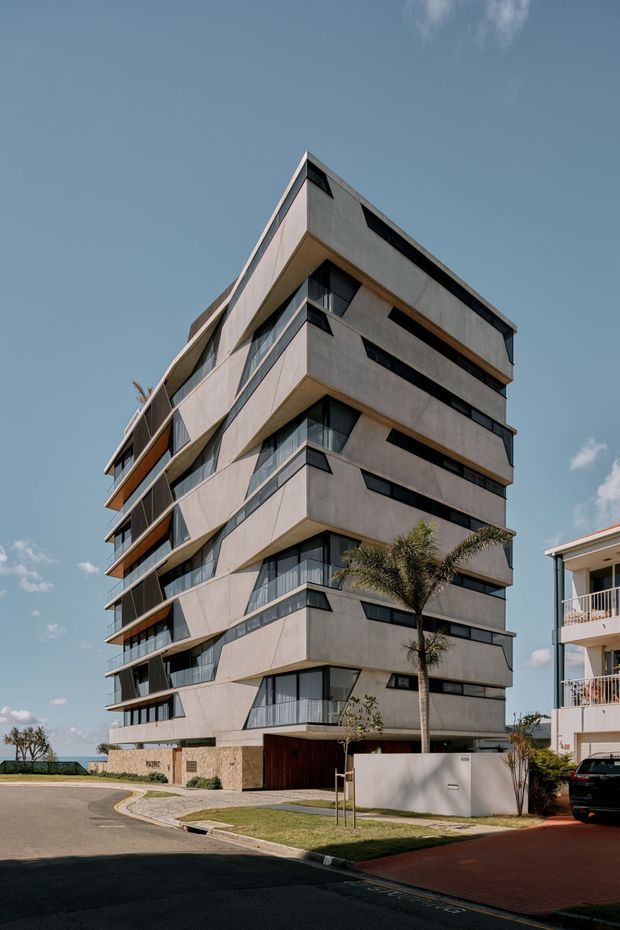 Street-level exterior photograph of a contemporary multi-storey apartment building with strongly cantilevered and staggered balconies, dark-framed glazing, pale concrete or rendered facade panels, and a flat roofline. The building stands on an urban residential street with landscaped frontage and neighboring apartments visible.