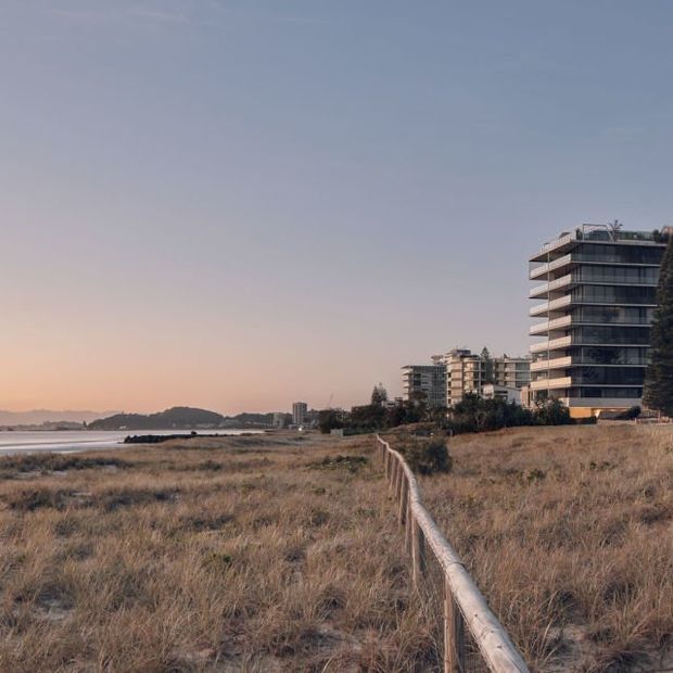 A coastal architectural exterior viewed across a dry grassy dune landscape, with a calm shoreline and distant hills on the left. Several contemporary multi-storey apartment buildings line the waterfront, including a prominent dark-glazed building with projecting horizontal balconies. A simple timber post-and-rail fence leads through the foreground toward the buildings. The image has warm low-angle evening light and a broad open sky.