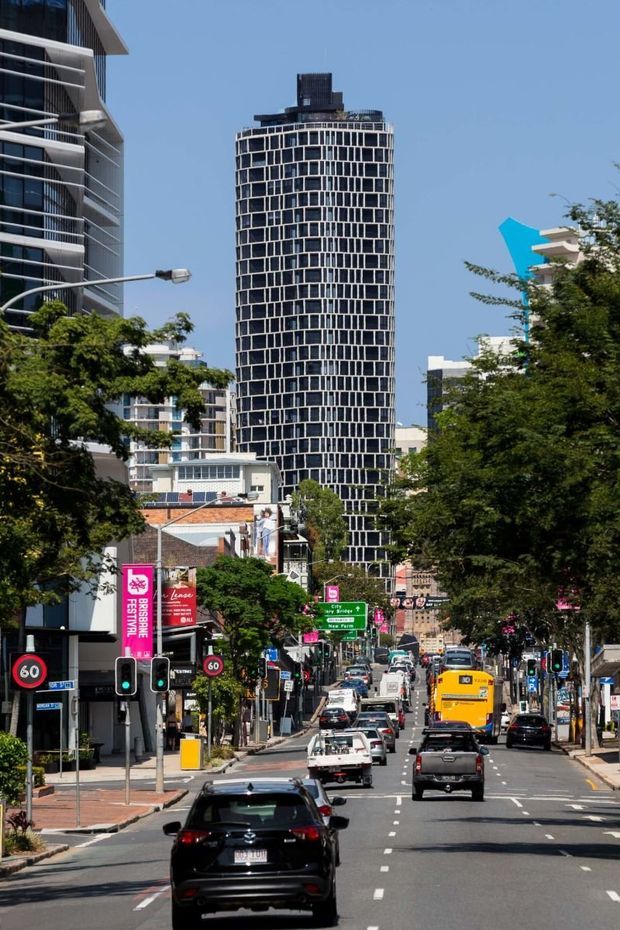 A bright daytime urban streetscape viewed along a busy city road, with a prominent tall cylindrical tower featuring a dense black-and-white rectangular window grid. Surrounding mid- and high-rise buildings, mature street trees, traffic signals, road signage, shops, and multiple vehicles frame the scene. The image is primarily an exterior architectural and urban context view.