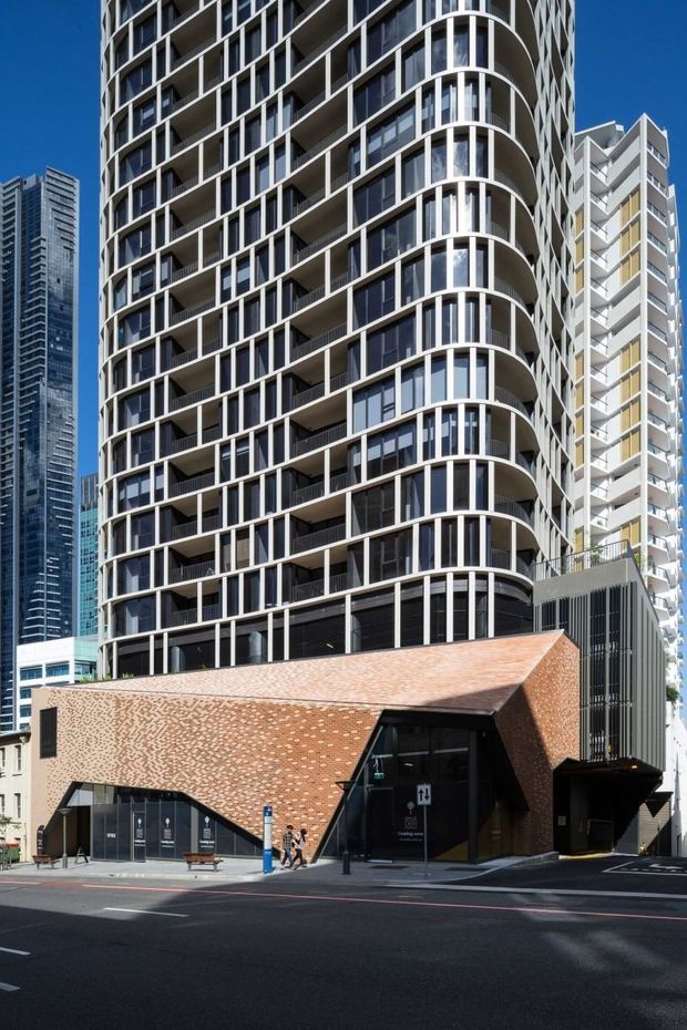 Street-level exterior view of a contemporary high-rise apartment or mixed-use building in a dense urban setting. The tower has curved corners, repetitive white structural framing, extensive dark glazing and balconies. A low angular brick-clad podium with a sharply pitched roofline and dark recessed entrance anchors the base, with neighbouring towers visible behind.
