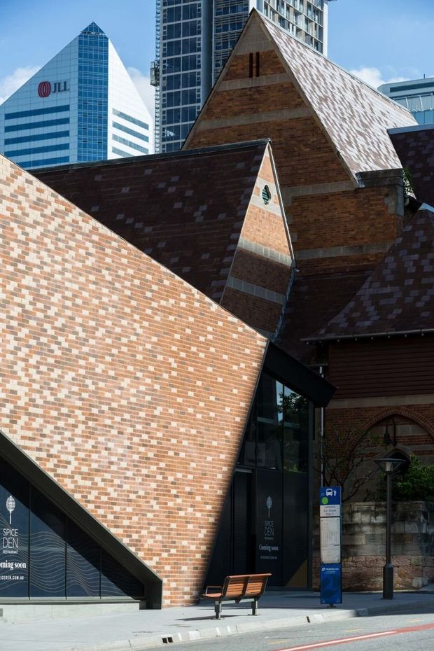 Urban exterior view of a distinctive brick building with steep triangular gabled roof forms, dark tiled roofing, and black-framed glazed entry doors. Modern high-rise office buildings are visible behind it, along with a street-level bench, signage, paving, and a bus stop.
