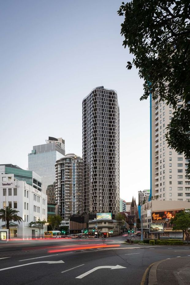 Street-level urban exterior photograph showing a dense city intersection with a prominent tall, rounded residential or commercial tower featuring a repetitive grid of balconies and windows. Surrounding mid- and high-rise buildings, retail frontages, street trees, traffic signals, and long-exposure vehicle light trails create a metropolitan streetscape.
