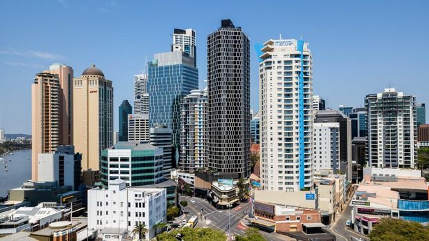 A wide daylight cityscape showing a dense cluster of modern high-rise commercial and residential buildings, with a waterfront visible at the left and roads and low-rise buildings in the foreground. The skyline features glass curtain walls, balconies, and varied contemporary towers under a clear blue sky.