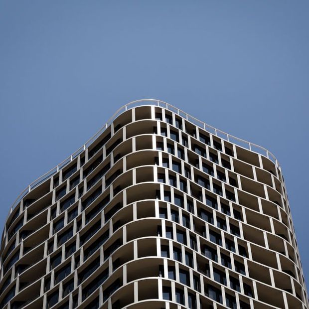 Low-angle exterior view of a contemporary multi-storey apartment building with rounded corners, extensive balconies, repetitive white structural framing, dark glazing, and a gently curved roofline against a clear blue sky.