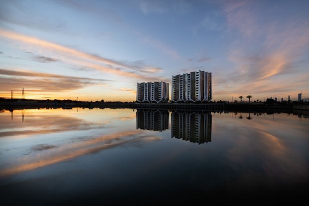 Wide exterior architectural photograph at sunset showing a cluster of tall modern apartment towers beside calm water, with strong reflections of the buildings and sky. The scene includes a flat waterfront setting, silhouetted palms, and distant infrastructure.
