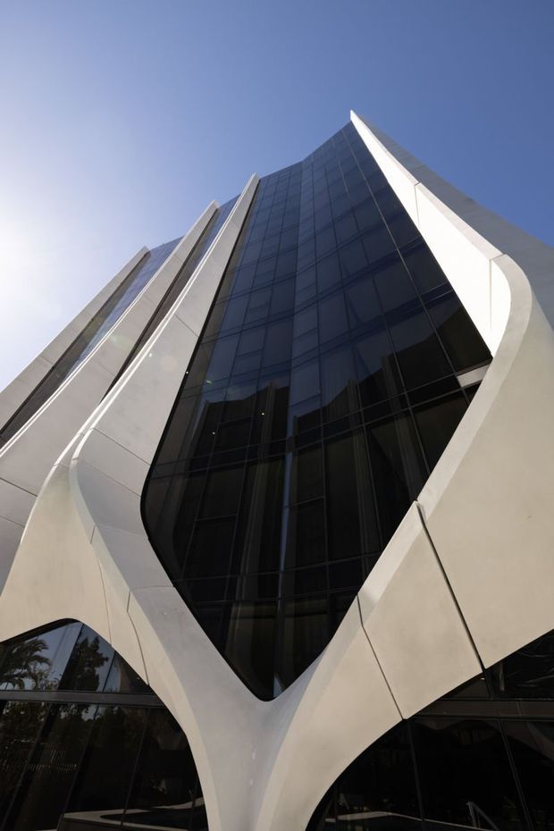 Low-angle exterior photograph of a tall contemporary commercial building. The facade features a dramatic dark glazed curtain wall framed by sweeping white sculptural structural elements that converge toward the pointed upper section. The reflective glazing and strong vertical geometry create a bold, futuristic appearance against a clear blue sky.