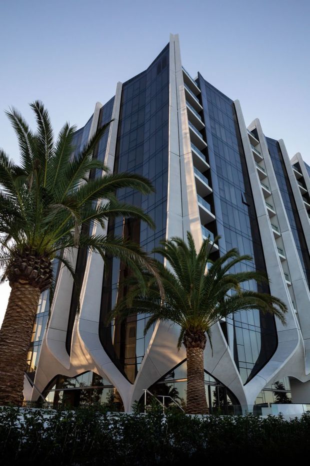Low-angle exterior view of a tall contemporary multi-storey commercial or mixed-use building. The facade combines extensive dark reflective glazing with pale sculptural structural fins and curved branching supports at the base, with projecting balconies along the sides. Palm trees and landscaped planting frame the entrance.