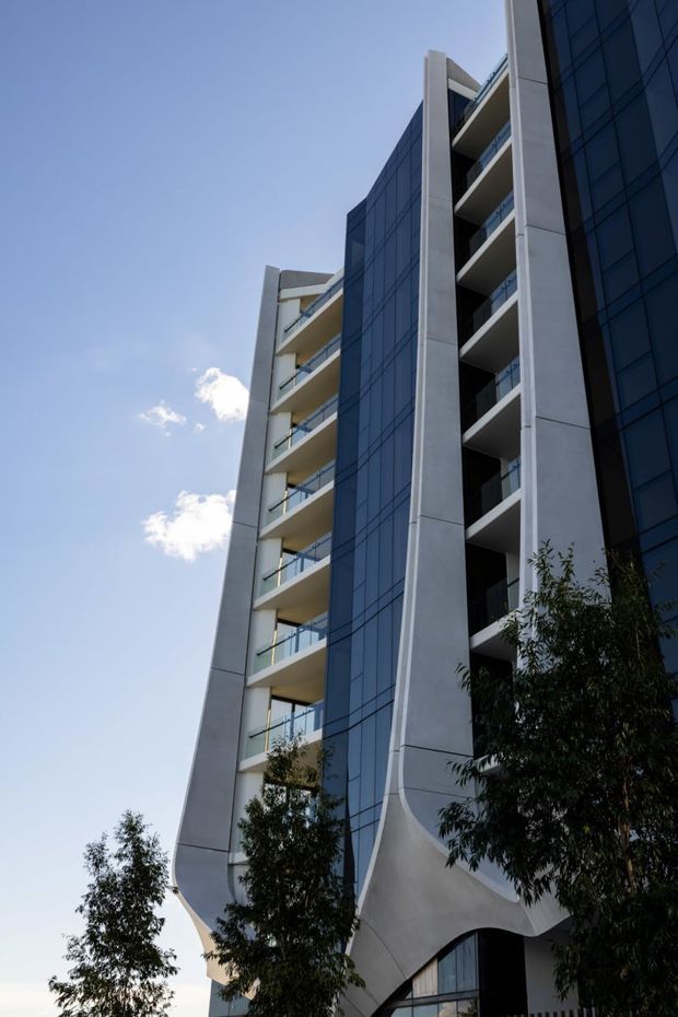 Low-angle exterior view of a contemporary multi-storey apartment or commercial building. The facade combines dark blue reflective curtain-wall glazing, pale concrete structural frames, recessed balconies with glass balustrades, and sculptural curved concrete forms at the lower level. Trees partially obscure the base, with a clear blue sky behind.