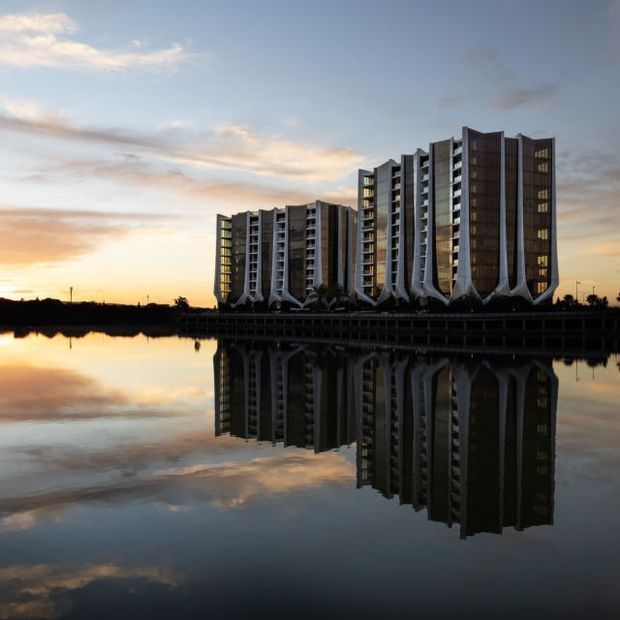 A pair of modern multi-storey residential towers beside calm water at sunset, with dramatic sculptural white structural elements at the base and a clear reflection in the water. The buildings feature dark vertical facade panels, extensive glazing, projecting balconies, and a contemporary high-rise form.