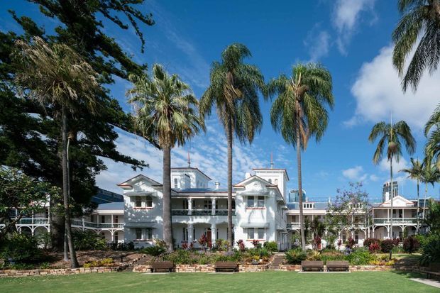A grand white multi-storey building with ornate verandas, balconies, pitched roof elements and classical detailing, set behind a landscaped lawn and framed by tall palm trees. The image appears to show a tropical or coastal resort-style exterior, with blue sky and distant urban elements visible beyond the property.