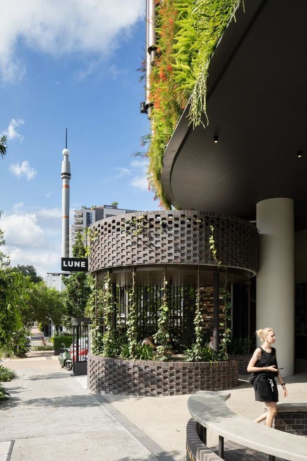 Street-level exterior view of a contemporary commercial building with a rounded brick screen enclosure, lush integrated planting, curved concrete bench seating, deep black soffits, and a rooftop garden. The scene includes a landscaped pedestrian walkway and a prominent LUNE sign.