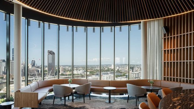 A sophisticated high-rise lounge or hospitality-style interior with expansive floor-to-ceiling glazing overlooking a dense city skyline. The room features a curved built-in upholstered banquette, freestanding lounge chairs and tables, a sculptural dark slatted ceiling with concealed perimeter lighting, sheer curtains, and a decorative timber screen wall. The palette is warm and refined, combining timber, cream upholstery, dark surfaces, and panoramic urban views.
