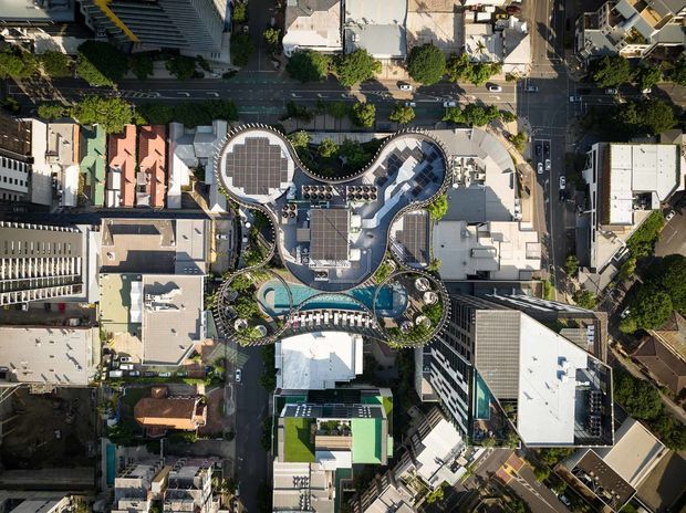 Aerial top-down view of a distinctive multi-storey urban building with curving, organic roof forms, extensive rooftop landscaping, terraces, and several rooftop swimming pools. The property is surrounded by dense city streets and neighbouring commercial and residential buildings.