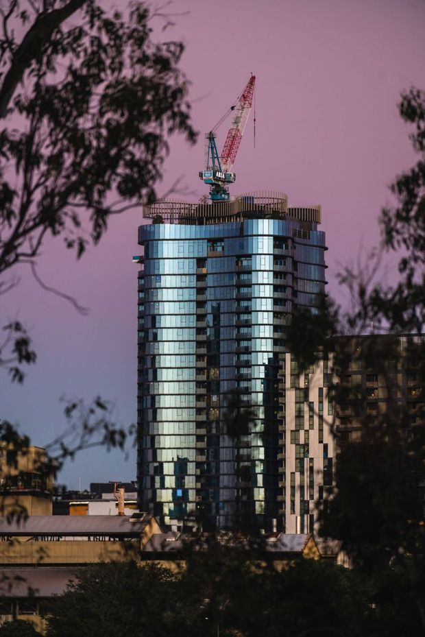 A cityscape view of a tall modern high-rise apartment or commercial building under construction, with curved glass curtain walls, dark vertical framing, and rounded rooftop elements. A tower crane is visible above the structure, while trees and lower-rise buildings frame the foreground. The scene is photographed in moody purple-pink dusk light.