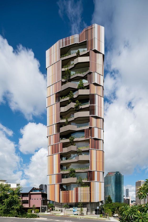 A tall contemporary multi-storey apartment or mixed-use tower viewed from the street, featuring a sculptural vertical form, alternating projecting balconies, extensive glazing, planted balcony edges, and a prominent facade of narrow vertical fins in varied warm and neutral tones. The building stands in an urban setting beneath a bright blue sky, with neighbouring high-rise buildings and street-level landscaping visible around it.