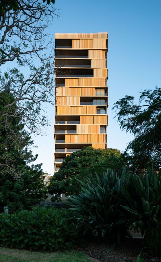 Tall contemporary multi-storey apartment building viewed externally through dense landscaped gardens. The narrow tower has a warm orange and golden vertical-panel facade, dark recessed balconies, and a simple flat roof, with extensive planting in the foreground and mature trees framing the building.
