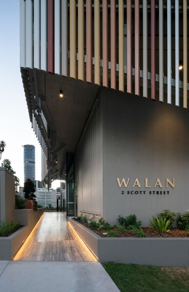 Photograph of a contemporary commercial building entrance at Walan, 2 Scott Street. The facade features a dark metal-clad wall with prominent vertical multicoloured fins, a deep projecting canopy with recessed lighting, illuminated signage, landscaped planter beds, and a polished stone or concrete pedestrian approach. A high-rise urban skyline is visible beyond the entry.