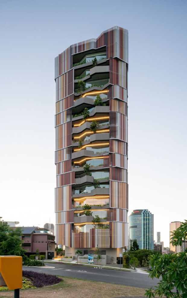 A tall contemporary multi-storey urban building with a narrow vertical form, screened in multicoloured vertical fins and articulated projecting balconies. Each level features planted terraces, dark slab edges, glass balustrades and warm linear lighting. The building stands in a landscaped suburban streetscape with other high-rise buildings visible in the background.