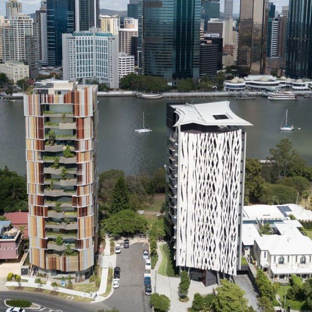 Aerial view of two distinctive modern high-rise buildings beside a waterfront, with a dense urban skyline across the water. One tower has colourful vertical facade panels and planted balconies, while the other features a bold white geometric screen facade and a sculptural roof. Landscaped grounds, access roads and neighbouring low-rise buildings are visible below.