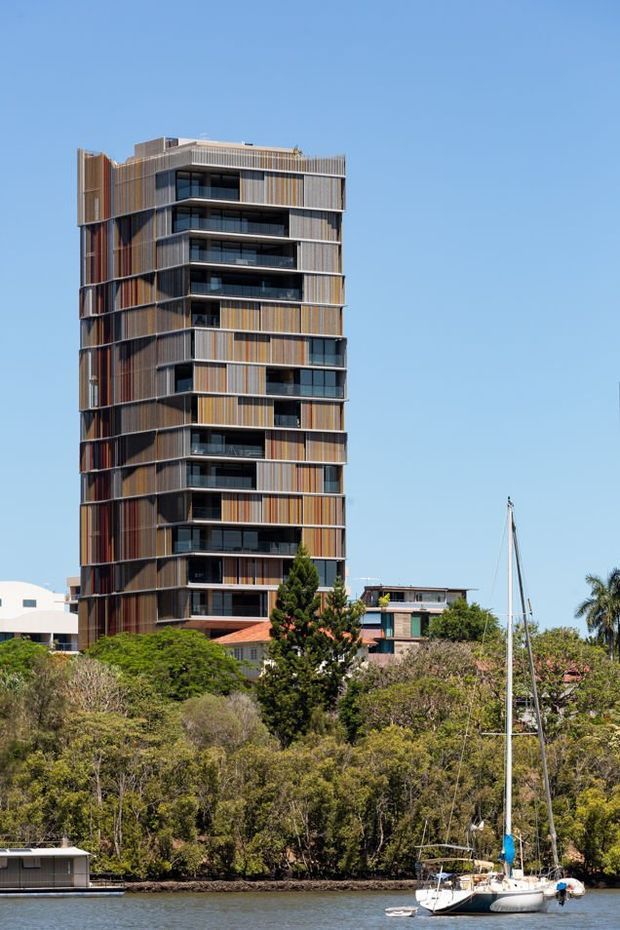 Exterior photograph of a tall contemporary multi-storey apartment building beside a wooded waterfront. The tower has a strongly articulated facade with alternating warm timber-toned vertical cladding, dark glazing, recessed balconies, and projecting horizontal floor bands. Trees, water, and a sailboat occupy the foreground.