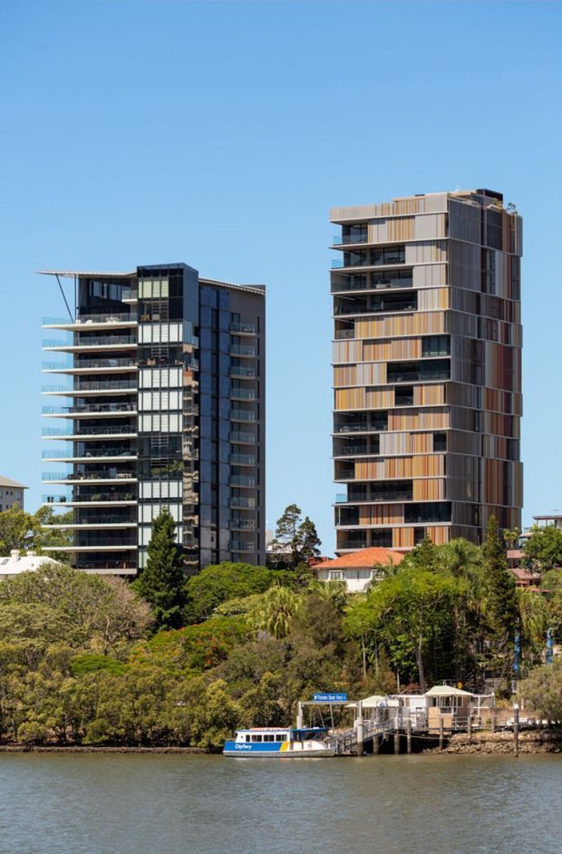 Exterior view across a river toward two tall contemporary apartment buildings, with balconies, extensive glazing, and vertically articulated cladding. Mature trees, low-rise homes, a waterfront ferry terminal, and clear blue sky are visible in the foreground and surroundings.