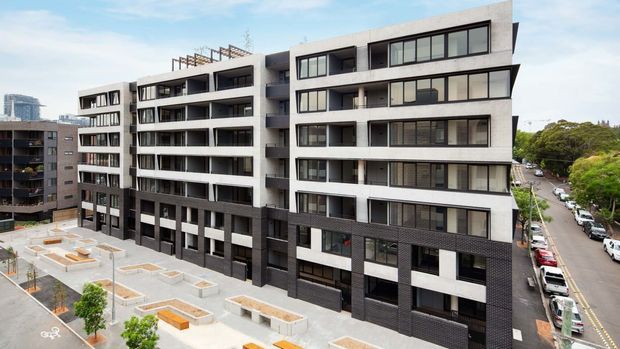 Exterior view of a contemporary multi-storey apartment building with contrasting light concrete and dark brick or masonry facade sections, extensive balconies, and large dark-framed windows. A landscaped communal courtyard and adjacent urban street are visible.