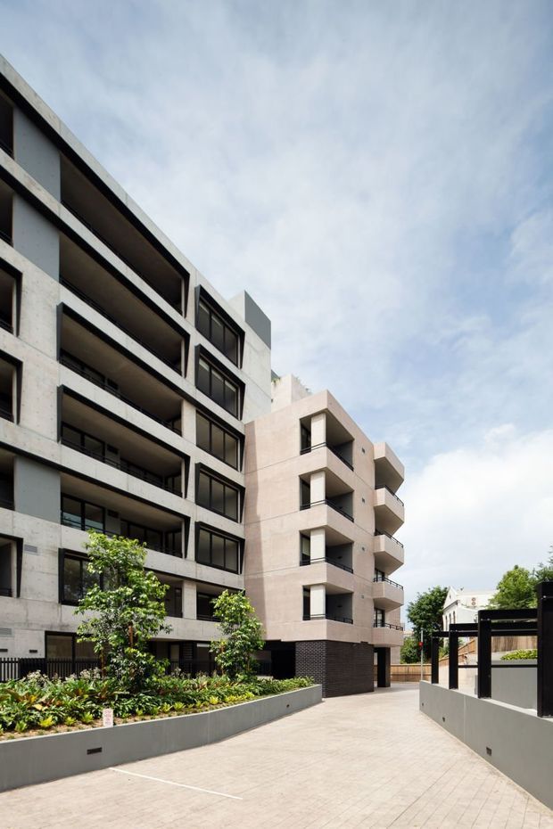 Contemporary multi-storey apartment building viewed from a landscaped paved driveway or courtyard. The facade combines exposed concrete, dark-framed glazing, recessed balconies, and a contrasting pale rendered wing, with low retaining planters and black metal fencing in the foreground under a bright overcast sky.