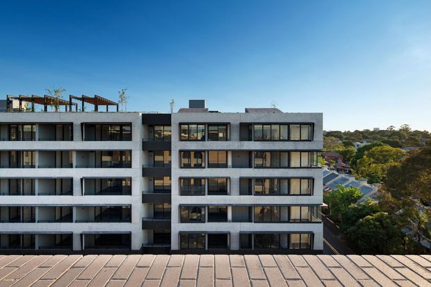 Exterior view of a contemporary multi-storey apartment building with a pale rendered facade, dark-framed floor-to-ceiling windows, projecting balconies, and a rooftop terrace structure. Residential suburban buildings and mature trees are visible in the background under a clear blue sky.