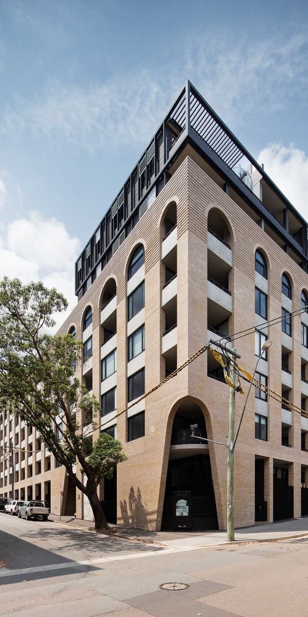 Street-level exterior photograph of a multi-storey urban apartment building. The facade combines pale tan brickwork with tall arched openings, dark-framed windows, recessed balconies, and a contemporary rooftop terrace enclosed by dark metal railings. Street trees, utility cables, pavements, and neighbouring buildings frame the view.