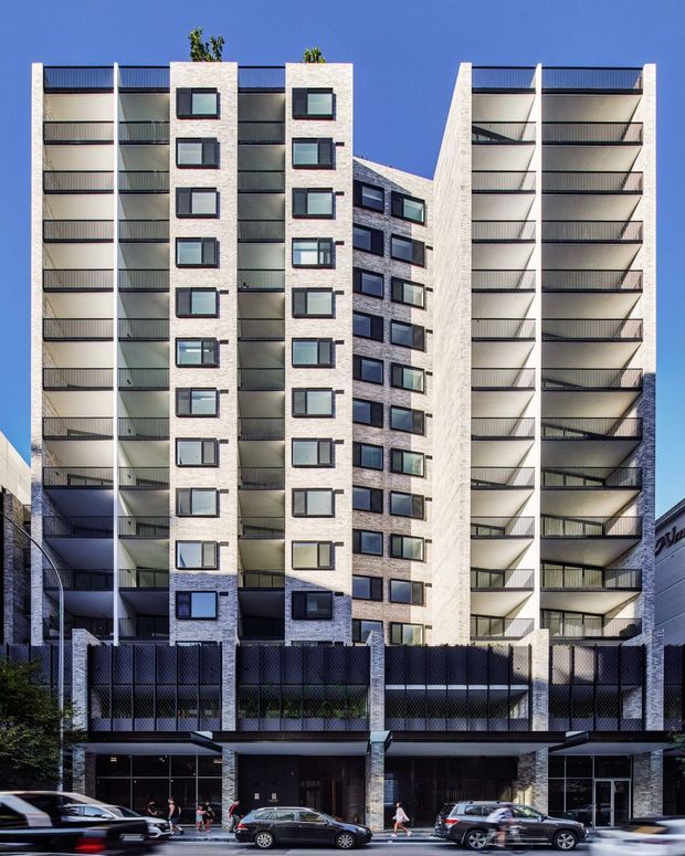 A tall contemporary multi-storey apartment building viewed from the street, with pale textured masonry walls, dark-framed windows, stacked recessed balconies, and a strongly symmetrical facade. The ground level contains glazed commercial or lobby-front spaces behind a dark metal fence, with mature street trees and parked vehicles in the foreground. The building rises against a clear blue sky.