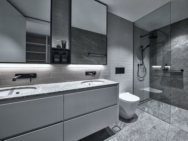 A contemporary grey bathroom featuring a long floating double vanity with integrated basins, pale grey subway-tile walls, dark stone-look shower surfaces, black fixtures, a frameless glass shower enclosure, and patterned grey floor tiles. Large mirrors and under-vanity lighting add brightness and visual depth.