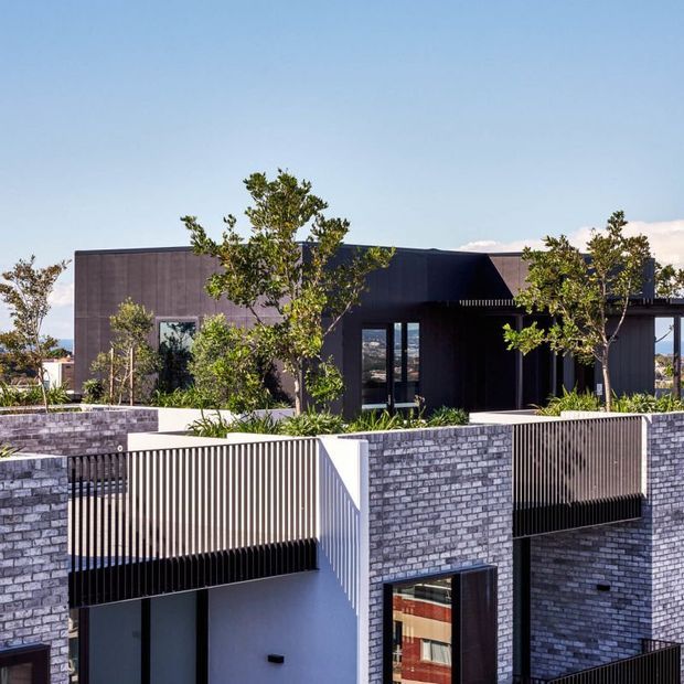 Contemporary multi-level residential building with dark cladding, flat roof forms, grey brick walls, vertical metal screening and landscaped terraces. The view is dominated by the exterior facade and shows strong geometric composition with glimpses of the surrounding landscape.