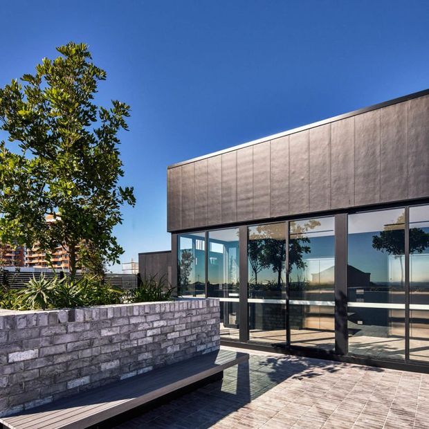 Contemporary flat-roof building exterior with a dark textured metal-clad upper facade, extensive floor-to-ceiling glazing, and a paved outdoor terrace. A low masonry planter wall with integrated bench seating borders landscaped planting, with an urban apartment building visible in the background.