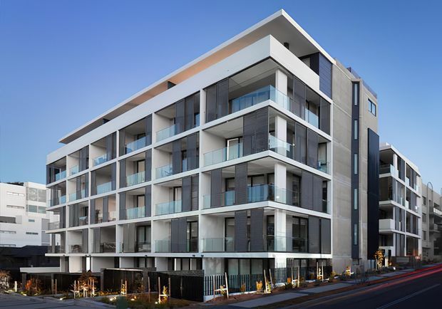 Exterior view of a contemporary multi-storey apartment building in an urban streetscape. The building has a strong white geometric frame, extensive floor-to-ceiling glazing, recessed balconies with clear glass balustrades, dark vertical cladding panels, and a flat roofline. Landscaped frontage and neighbouring buildings are visible.