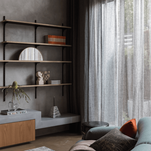 A moody contemporary living room corner with open black metal shelving, a concrete-look built-in console, timber cabinetry, decorative objects, and a large window dressed with layered sheer and dark curtains. A blue-grey sofa with rust and dark cushions occupies the foreground.