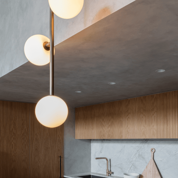A sophisticated contemporary kitchen or kitchenette interior featuring a dark textured ceiling, warm vertical timber slat wall panelling and cabinetry, pale stone or concrete-effect wall surfaces, and a sculptural globe pendant light with brass-toned fittings. A compact sink and faucet are visible beneath the timber cabinetry, with recessed ceiling lights adding subtle illumination.