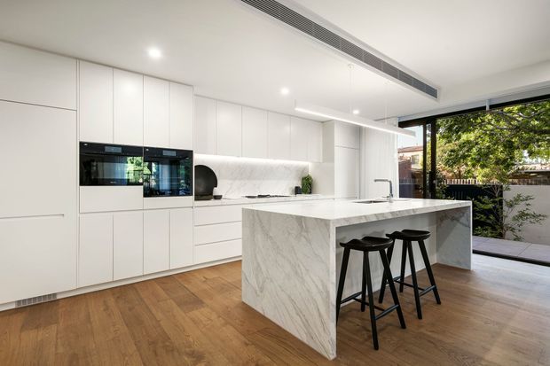 Bright contemporary kitchen featuring extensive white handleless cabinetry, integrated black appliances, a large white marble-look island with waterfall sides and an undermount sink, and two black bar stools. Warm timber flooring contrasts with the predominantly white scheme. Full-height black-framed glazing opens visually to a landscaped outdoor area, while recessed lighting and a linear ceiling air-conditioning vent create a clean, refined finish.